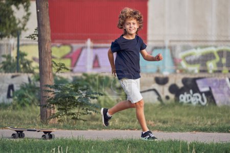 Foto de Vista lateral de niño alegre con el pelo rizado en ropa casual y zapatillas de deporte corriendo a lo largo de la acera cerca de monopatín mientras mira a la cámara - Imagen libre de derechos