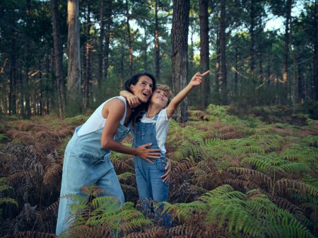 Photo for Woman with mouth opened and smiling boy pointing away in matching outfits hugging while walking among ferns and tall spruce trees - Royalty Free Image