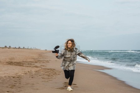 Foto de Cuerpo completo de alegre joven blogger con cálida camisa a cuadros sonriendo y corriendo a lo largo de la playa de arena mientras graba vlog en la cámara contra el cielo nublado - Imagen libre de derechos