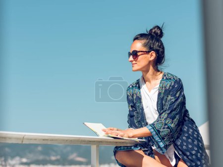 Foto de Vista lateral de la hermosa joven hembra en traje elegante y gafas de sol sentado con libro cerca de barandilla en el balcón mientras mira hacia otro lado en el soleado día de verano - Imagen libre de derechos