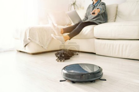 Photo for Crop unrecognizable person in casual clothes sitting with laptop on knees on sofa near sleeping dog and using remote control on robotic vacuum cleaner - Royalty Free Image