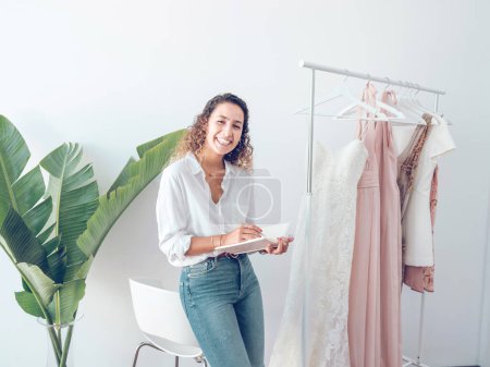 Téléchargez les photos : Joyeux designer féminin en chemisier et jeans debout avec ordinateur portable dans les mains près du rack avec des robes élégantes dans la salle de lumière - en image libre de droit