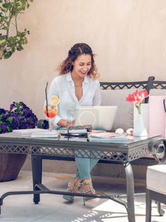 Foto de Cuerpo completo de mujer freelancer positivo sentado en el sofá en la terraza de la cafetería y ordenador portátil de navegación con cóctel fresco en la mesa a la luz del sol - Imagen libre de derechos