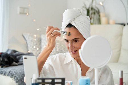Photo for Focused adult smiling female influencer with towel on head applying foundation on forehead using brush while recording online tutorial for makeup blog at home - Royalty Free Image