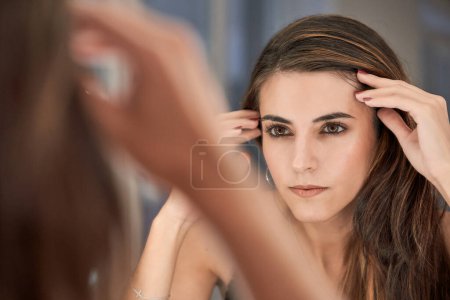 Téléchargez les photos : Charmant modèle jeune femme avec maquillage touchant la tête et regardant reflet dans le miroir tout en étant assis dans un salon de beauté moderne - en image libre de droit