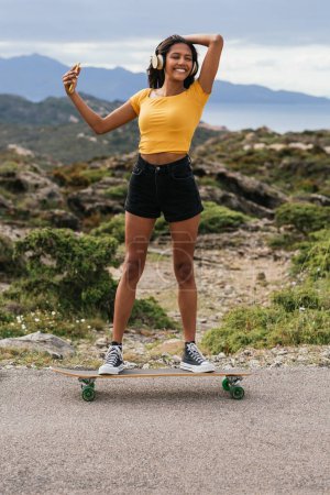 Téléchargez les photos : Ethnique femelle prendre selfie sur smartphone tout en se tenant sur longboard sur route asphaltée - en image libre de droit