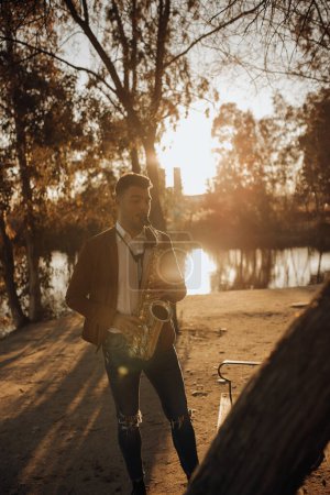 Photo for Young romantic musician playing on saxophone in park near water in sunny day - Royalty Free Image