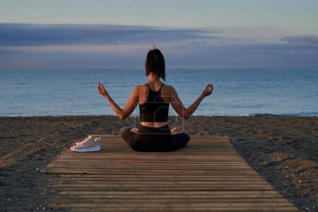 Rückansicht einer anonymen Frau, die in Lotus-Pose mit Mudra-Händen sitzt, während sie abends am Strand am Meer meditiert