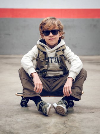 Photo for Full body of serious boy in stylish clothes and sunglasses sitting on skateboard on concrete floor while looking at camera - Royalty Free Image