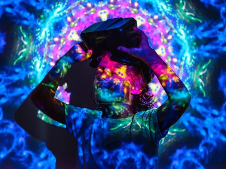 Photo for Dreamy boy with closed eyes wearing VR headset showing OK gesture while standing under bright blue neon abstract projector light - Royalty Free Image