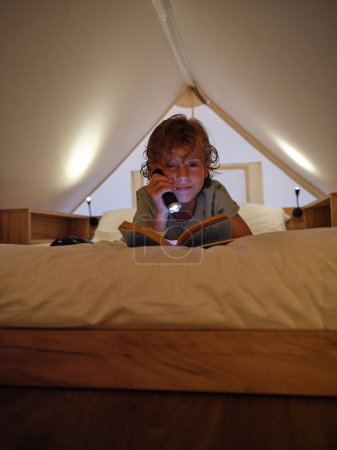 Téléchargez les photos : Contenu garçon brillant lampe de poche au livre et en regardant la caméra tout en étant couché sur un lit blanc confortable sur mansarde à la maison - en image libre de droit