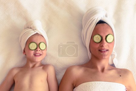 Photo for Top view of relaxed female and kid with towels on heads and cucumber slices on eyes resting on white bed - Royalty Free Image