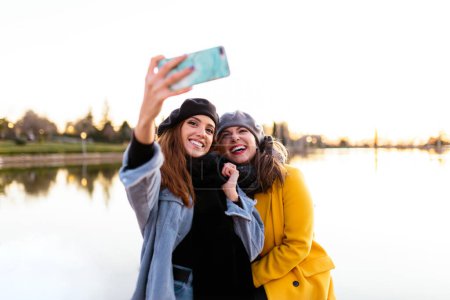 Téléchargez les photos : Femmes occasionnelles prenant selfie sur smartphone au lac ensoleillé - en image libre de droit