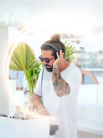 Téléchargez les photos : Concentré tatoué Hispanique gars écouter de la musique dans les écouteurs tout en se tenant sur la terrasse et de travailler avec console DJ en été - en image libre de droit
