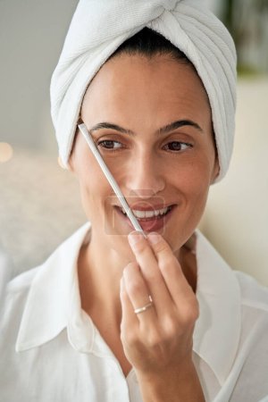 Téléchargez les photos : Femme adulte positive avec serviette sur la tête appliquant une brosse cosmétique près des sourcils tout en faisant du maquillage et en vérifiant la forme parfaite des sourcils et en détournant les yeux - en image libre de droit