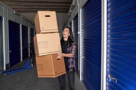 Positive weibliche Blicke beim Tragen eines Stapels Kartons mit persönlichen Gegenständen in der Nähe des geschlossenen Selfstorage während des Umzugstages
