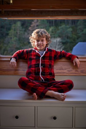 Photo for Cheerful boy with blond curly hair in red pajama sitting on white cabinet and leaning on opened window in apartment in daytime - Royalty Free Image