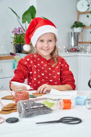 Photo for Positive girl in red Santa hat applying white icing on baked gingerbread cookies while looking at camera during Christmas preparation - Royalty Free Image
