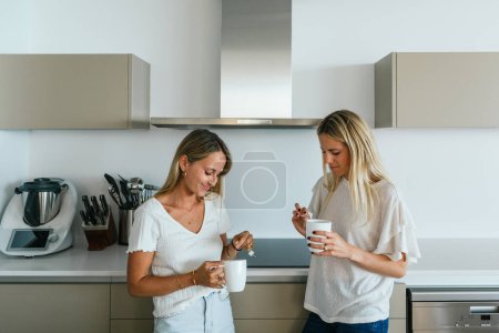 Téléchargez les photos : Amis féminins positifs avec des tasses de café en se regardant tout en se tenant dans la cuisine avec des ustensiles de cuisine modernes à la maison - en image libre de droit