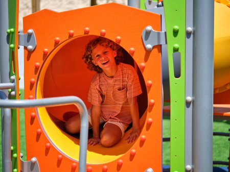Photo for Adorable little kid sitting in colorful tube slide while smiling happily and looking at camera - Royalty Free Image