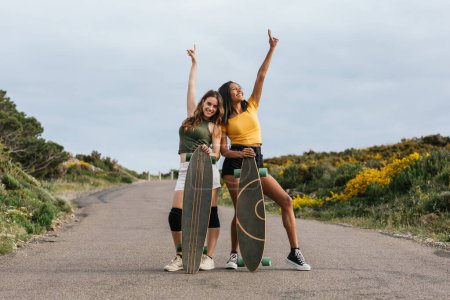 Foto de Cuerpo completo de amigas multiétnicas felices con brazos extendidos de pie en la carretera con longboards - Imagen libre de derechos