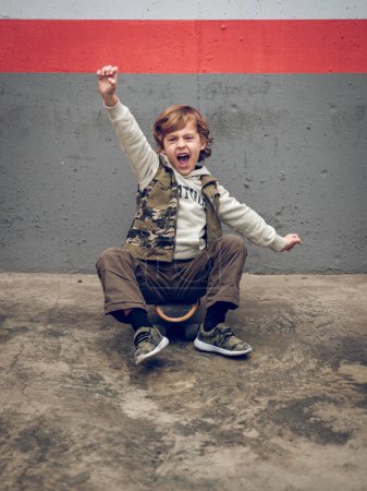 Photo for Full body of excited boy in trendy clothes sitting with raised arms and open mouth on skateboard on street - Royalty Free Image