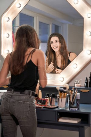 Foto de Mujer delgada positiva con maquillaje mirando a la reflexión en el espejo con bombillas mientras toca el pelo largo y oscuro - Imagen libre de derechos