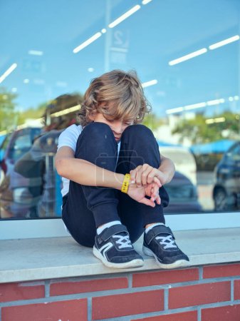 Foto de Retrato de un niño solitario sentado cerca de una ventana con coches aparcados reflejándose en ella y abrazando las rodillas mientras se siente molesto - Imagen libre de derechos