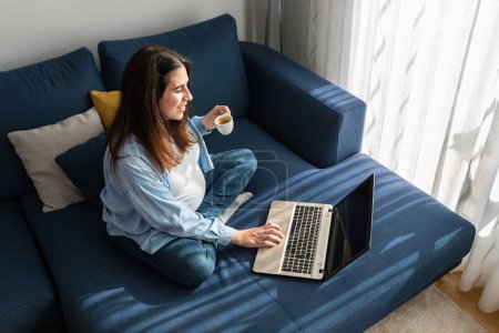 Téléchargez les photos : Femme enceinte positive assise sur un canapé doux avec ordinateur portable et boire une boisson chaude - en image libre de droit