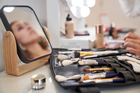 Téléchargez les photos : Gros plan de assortiment de divers pinceaux de maquillage professionnels en mallette en cuir et miroir avec réflexion de la femme placée sur la table dans un studio de beauté - en image libre de droit
