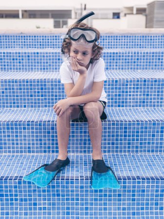 Téléchargez les photos : Corps complet de triste garçon préadolescent bouleversé en masque de plongée avec tuba et palmes assis sur les marches de la piscine vide - en image libre de droit