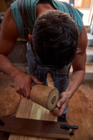 Foto de Trabajador de madera masculino calificado sin rostro en detalles de madera tallada en ropa de trabajo con cincel especial y mazo mientras trabaja en un taller profesional ligero - Imagen libre de derechos