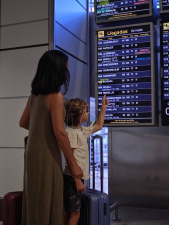Foto de Vista trasera del niño anónimo con ropa casual señalando el horario de salida y la mujer sosteniendo la maleta en la terminal del aeropuerto que se va de vacaciones durante las vacaciones de verano - Imagen libre de derechos