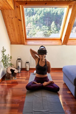 Foto de Cuerpo completo de hembra descalza en gafas de realidad virtual y sentada en pose de Loto con piernas cruzadas mientras practica meditación de yoga en casa - Imagen libre de derechos