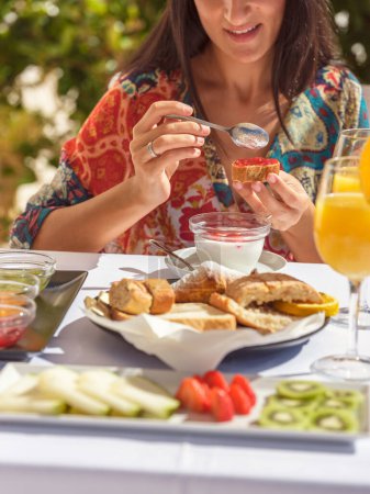 Foto de Recorte viajero femenino irreconocible aplicación de mermelada en tostadas en la mesa con una variedad de alimentos frescos durante el desayuno en la terraza del hotel soleado - Imagen libre de derechos