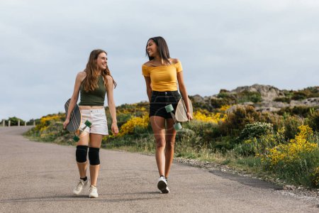 Foto de Cuerpo completo de amigas multiétnicas felices mirándose mientras caminan por la carretera con longboards - Imagen libre de derechos