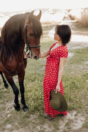 Foto de Joven hembra en vestido rojo largo con caballo mientras está de pie en el patio del rancho en el día soleado en el campo - Imagen libre de derechos