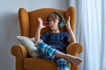 Foto de Niño en auriculares sentado en sillón con la tableta en las rodillas y jugando con la cuerda de dedo de colores pasar el fin de semana en casa - Imagen libre de derechos