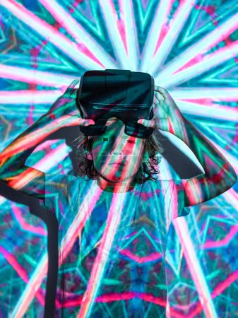Téléchargez les photos : Le gamin enlève ses lunettes VR après s'être immergé dans le monde virtuel tout en se tenant sous un néon brillant illuminant un projecteur abstrait bleu brillant et en regardant une caméra - en image libre de droit