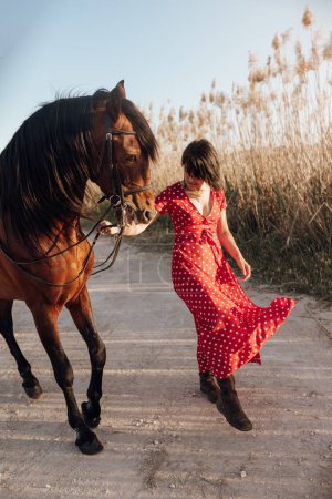 Téléchargez les photos : Dame en robe rouge tachetée menant cheval de prairie sur le chemin près de prairie - en image libre de droit