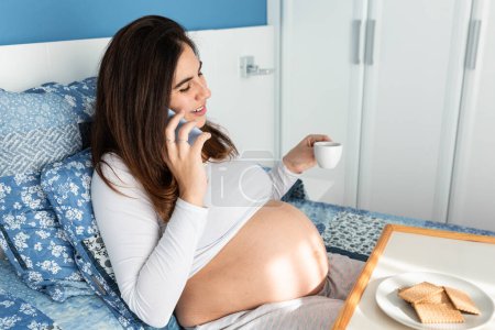 Foto de Vista lateral de la alegre hembra embarazada sentada en la cama por la mañana y desayunando mientras habla en el teléfono inteligente - Imagen libre de derechos
