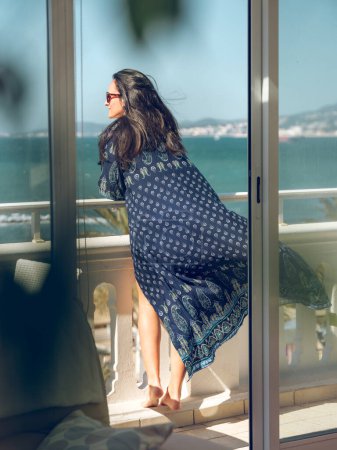 Foto de A través de la vista trasera de cristal de la mujer en ropa de verano y gafas de sol apoyadas en la barandilla de piedra de la terraza en el hotel mientras mira hacia otro lado en un día soleado con el océano en el fondo - Imagen libre de derechos