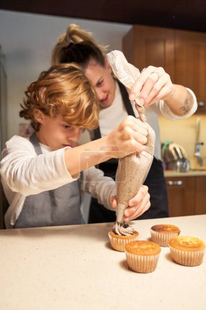 Téléchargez les photos : Photo verticale d'une fille portant un tablier et aidant un garçon à décorer des petits gâteaux avec un sac à pâtisserie - en image libre de droit