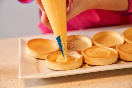 Foto de Cosecha anónima hembra pastelera haciendo picos de crema en tartaletas mientras está de pie en la cocina de la panadería casera - Imagen libre de derechos