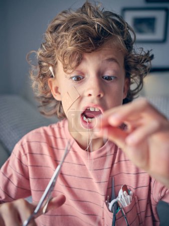 Foto de Niño asustado en camiseta con hisopos de algodón cortando hilo en diente de leche con tijeras mientras está de pie en la sala de luz - Imagen libre de derechos