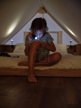 Photo for Full barefoot focused barefoot boy reading interesting book with flashlight while sitting on comfortable bed in mansard with dim light - Royalty Free Image