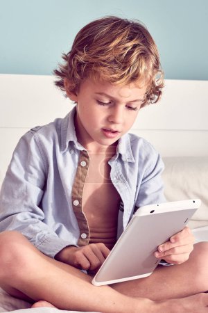 Photo for Cute boy wearing unbuttoned shirt sitting with crossed legs and playing video game on modern digital tablet in bedroom - Royalty Free Image