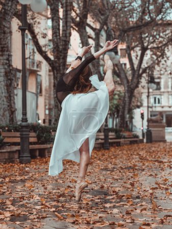 Photo for Side view of charming unrecognizable ballerina in elegant white skirt doing standing split on street in autumn on blurred background - Royalty Free Image