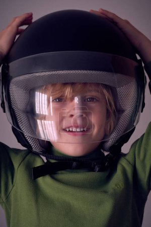 Photo for Cute little kid wearing big black protective helmet with transparent plastic front covering face ad touching head raising hands while looking at camera - Royalty Free Image