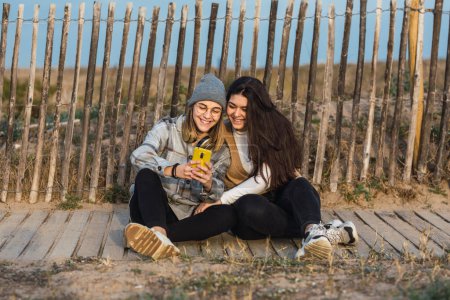 Photo for Full body of young lady with long dark hair in casual clothes sitting on wooden path in rural terrain and hugging female best friend using smartphone - Royalty Free Image